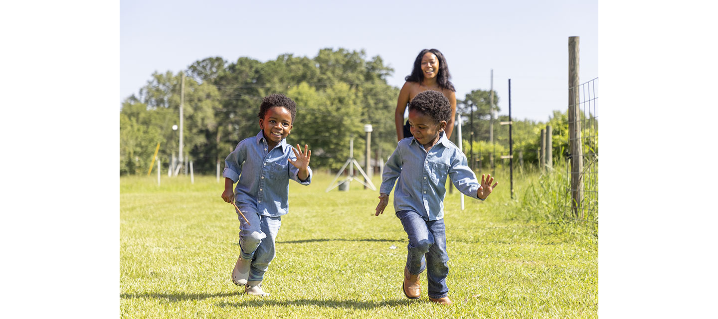 Twin boys playing with their mom, running in a grassy field with trees behind them.